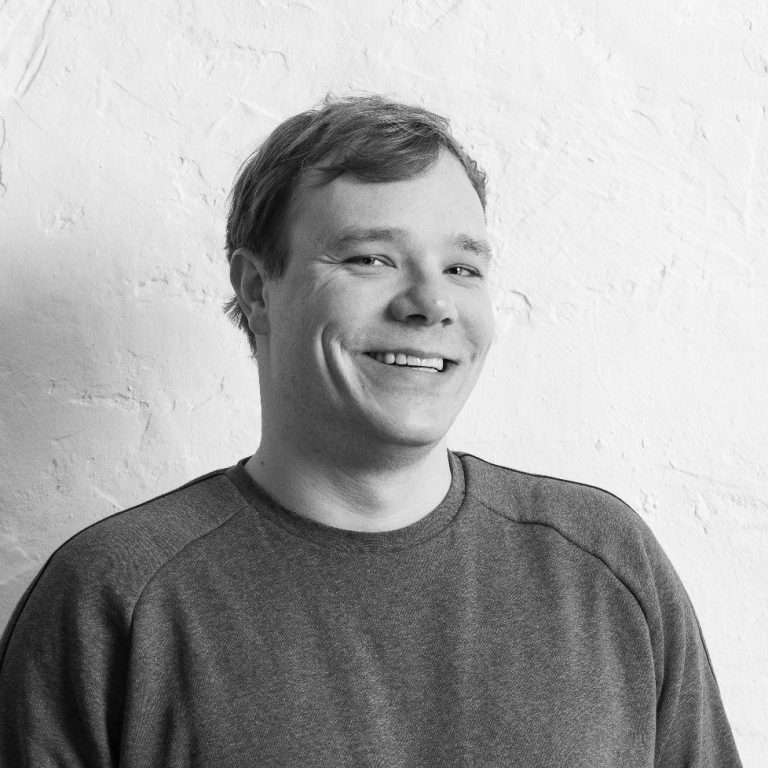 Felix Franzke lächelnd in einem grauen Pullover steht vor einer weißen Wand.