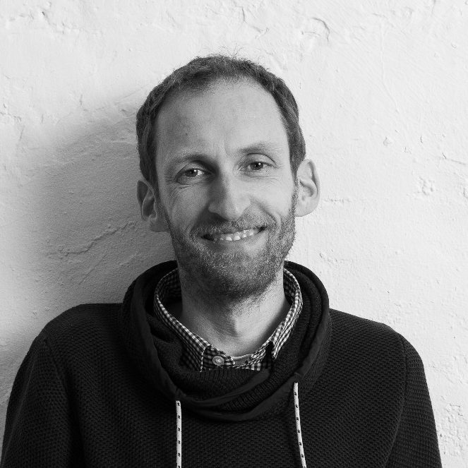 Björn Hinrichs mit kurzer Haarfrisur, Lächeln, schwarze Kapuzenjacke und hellem Hintergrund.