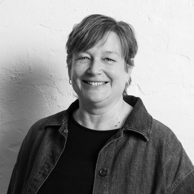 Miriam Schulz lächelnd mit kurzen Haaren, trägt einen schwarzen Oberteil. Heller Hintergrund.