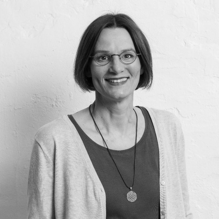 Christiane Brinkmann lächelnd mit kurzem Haar, Brille und beigem Cardigan vor einer hellen Wand.