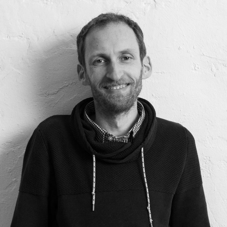 Björn Hinrichs Schulsozialarbeiter Schwarz-Weiß-Foto von Schulsozialarbeiter Björn Hinrichs. Lächelnd steht er vor einer hellen, strukturierten Wand.