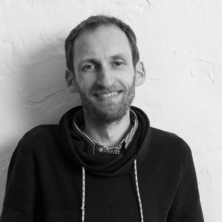 Schwarz-Weiß-Foto von Schulsozialarbeiter Björn Hinrichs. Lächelnd steht er vor einer hellen, strukturierten Wand.