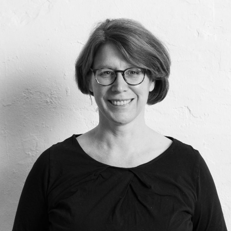 Barbara Ellerhorst, lächelnd mit Brille steht vor einer weißen Wand. Sie trägt ein schwarzes Oberteil.