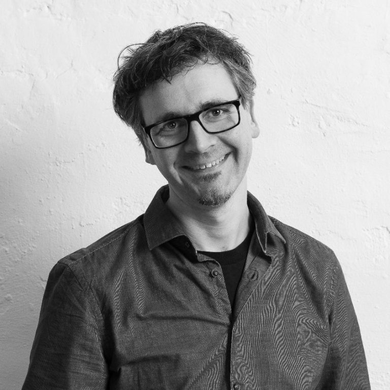 Kristof Nieroba lächelnd mit Brille in einem grauen Hemd vor einer neutralen Wand.