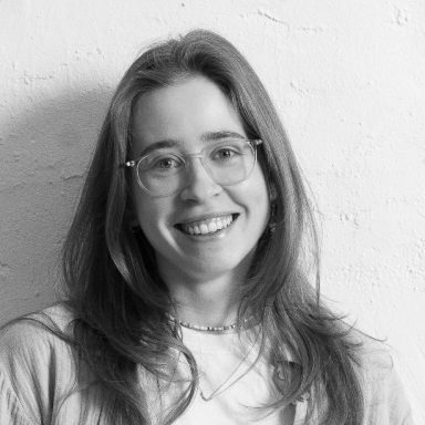 Nicole Artz mit langen Haaren und Brille, lächelt vor einer hellen Wand.