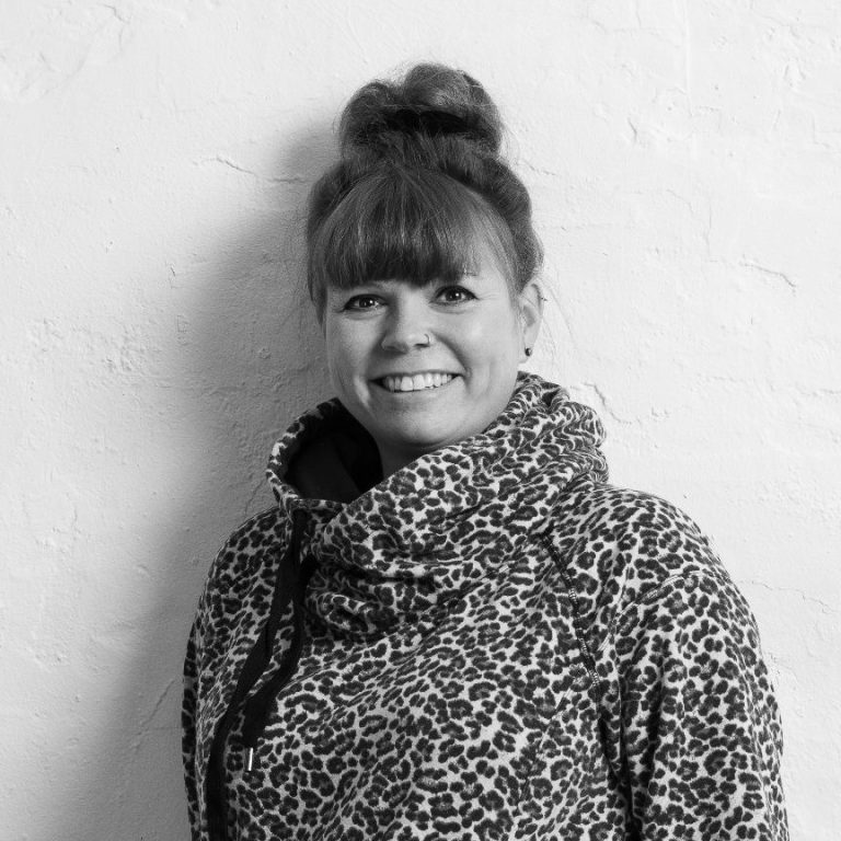 Kristina Kretschmer Schulsozialarbeiterin Schwarz-Weiß-Foto von Schulsozialarbeiterin Kristina Kretschmer, lächelnd in einem gemusterten Hoodie steht vor einer hellen Wand.