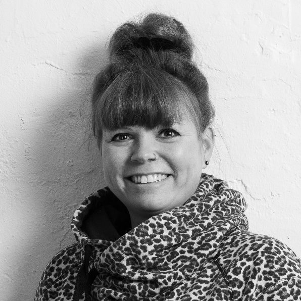Schwarz-Weiß-Foto von Schulsozialarbeiterin Kristina Kretschmer, lächelnd in einem gemusterten Hoodie steht vor einer hellen Wand.