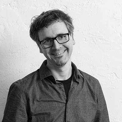 Kristof Nieroba Kristof Nieroba lächelnd mit Brille in einem grauen Hemd vor einer neutralen Wand.