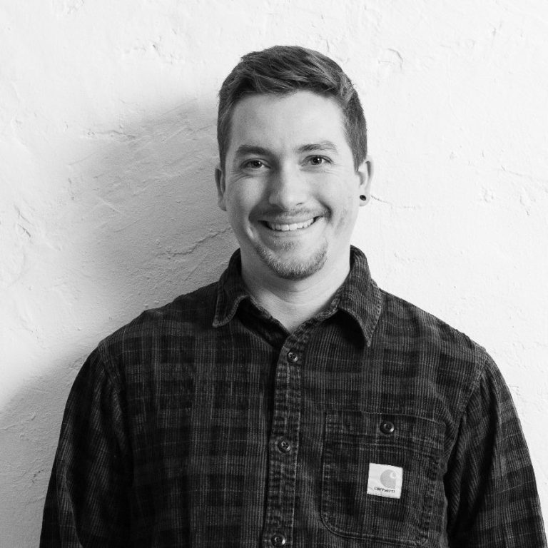 Till Bley steht lächelnd vor einer hellen Wand, trägt kariertes Hemd und Jeans.