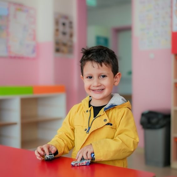 Ein lächelnder Junge in gelbem Mantel spielt mit Spielzeugautos in einem Klassenzimmer.