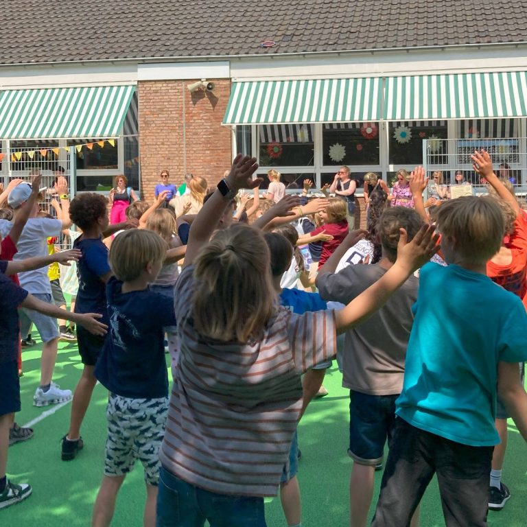 Eine Gruppe von Kindern tanzt und hebt die Hände auf einem grünen Platz.