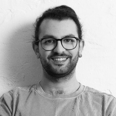Mohammad Dahman lächelnd mit Brille und lockigem Haar, stehend vor einer hellen Wand.