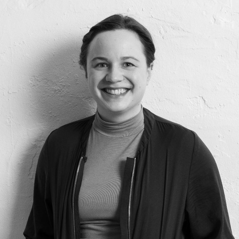 Sophie Minne mit zusammen gebundenem Haar, lächelt, steht vor einer hellen Wand.