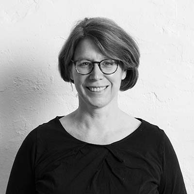 Barbara Ellerhorst Barbara Ellerhorst, lächelnd mit Brille steht vor einer weißen Wand. Sie trägt ein schwarzes Oberteil.