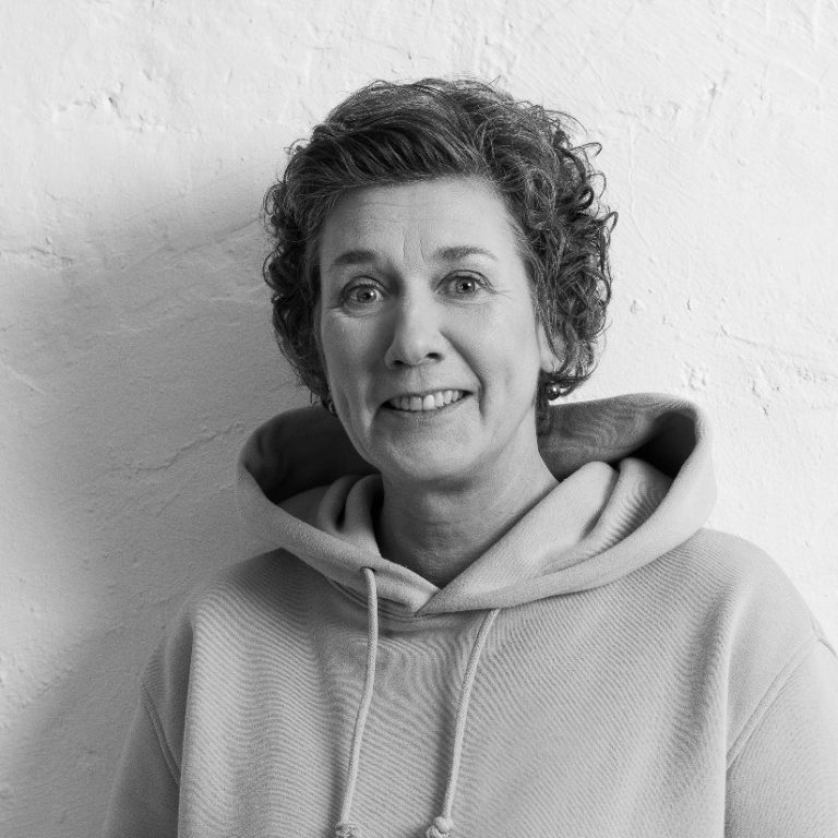 Ulrike Junker, lächelnd in einem grauen Hoodie, stehend vor einer hellen Wand.