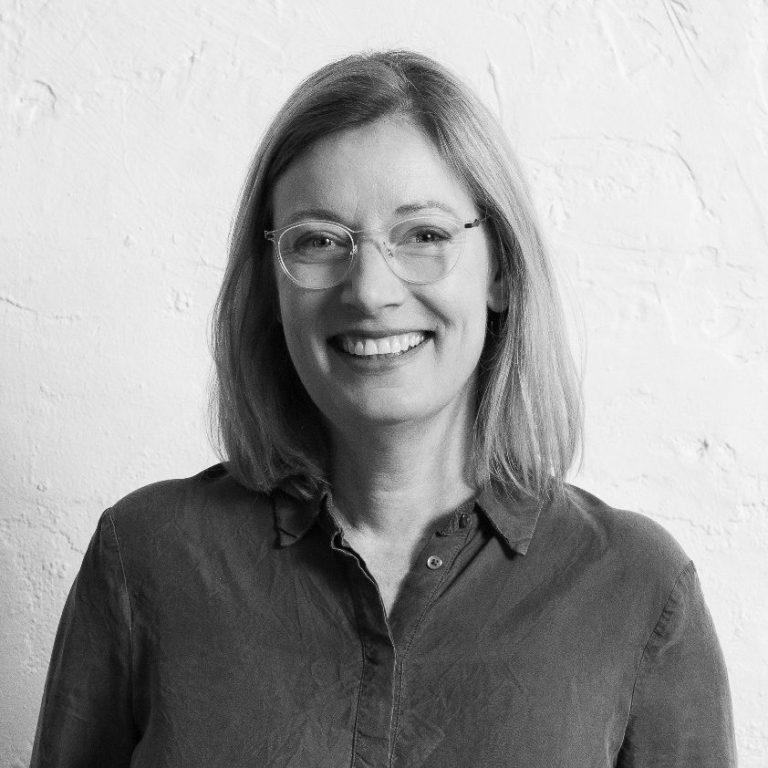 Katrin Schuppelius Katrin Schuppelius mit blonden Haaren und Brille, lächelt vor hellem, strukturiertem Hintergrund.