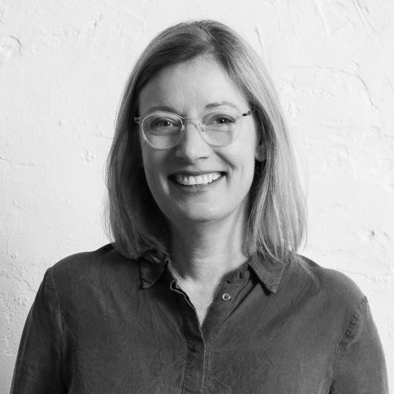 Katrin Schuppelius mit Brille und blonden Haaren vor einem hellen, strukturierten Hintergrund.
