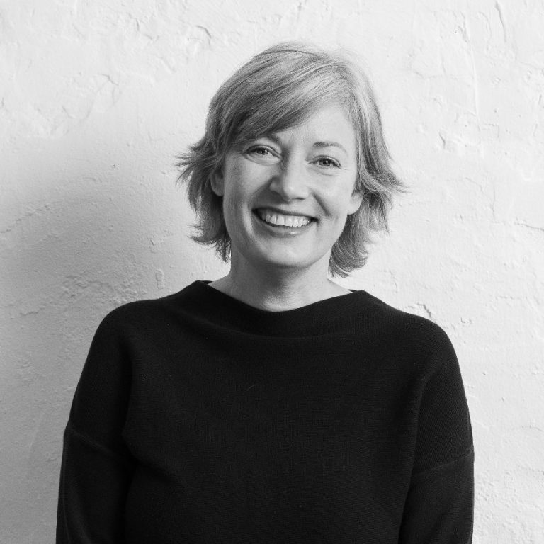 Katrin Schuppelius lächelnd in einem schwarzen Pullover vor einer hellen Wand mit kurzen blonden Haaren.