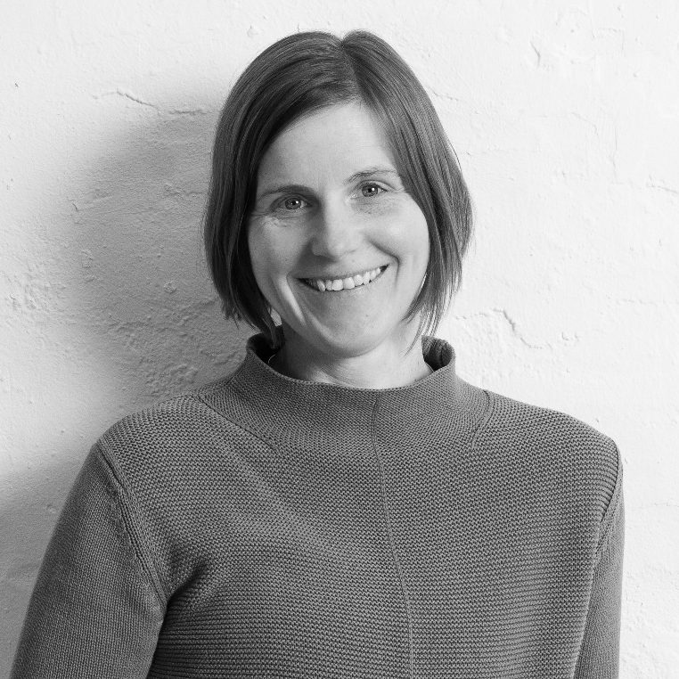 Thea Berthold-Bastrop Thea Berthold-Bastrop in einem grauen Pullover steht lächelnd vor einer weißen Wand.